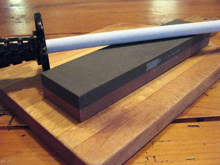 A white ceramic honing rod resting on a two-sided sharpening stone, on a wooden board